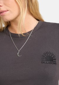 Camiseta de rayas gris oscuro con un logo de sol y el nombre de la marca bordado en negro, acompañada de un collar de media luna plateada.