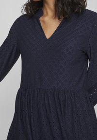Mörkblå klänning med V-ringning, långa ärmar och samlad midja. Tyget har strukturerade diamantmönster med hålbroderier.