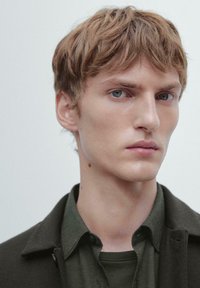 Ritratto di una modella con capelli corti, spettinati e castano chiaro, che indossa una giacca verde scuro sopra una camicia verde, su uno sfondo neutro.