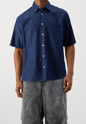 Homme portant une chemise à manches courtes bleu foncé boutonnée, portée en dehors d'un jean gris délavé ample sur un fond uni.