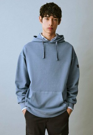 Felpa blu chiaro in cotone, con tasca frontale, cappuccio regolabile con stringhe e polsini a coste, indossata sopra una camicia bianca con colletto.
