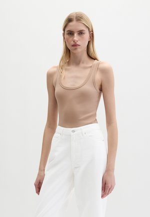 Femme portant un débardeur côtelé beige et un pantalon blanc taille haute, debout devant un fond clair uni.