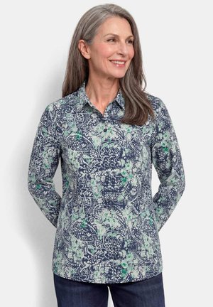 Smiling middle-aged woman with long gray hair wearing a blue and green patterned long-sleeve shirt with collar, standing against a plain background.