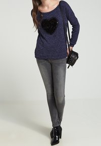 Marinblå tröja med en stor svart paljetthjärtform, bärs tillsammans med grå tajta jeans och svarta ankla stövlar, kompletterat med en axelväska.