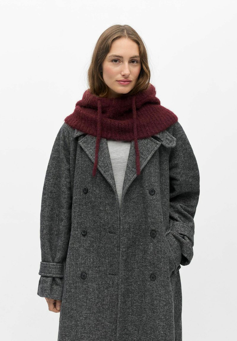 Femme portant un manteau gris croisé à double boutonnage et une grande écharpe en maille bordeaux avec des cordons, debout devant un fond blanc uni.