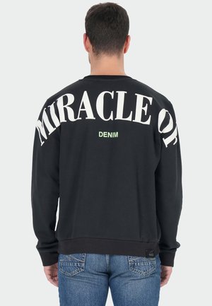 Man draagt een zwarte sweat met grote witte tekst "MIRACLE OF" en kleinere groene tekst "DENIM" op de rug, gecombineerd met blauwe spijkerbroek.