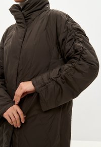 Persona che indossa una giacca imbottita marrone scuro con maniche arricciate, mentre chiude una cerniera frontale nascosta su uno sfondo neutro.