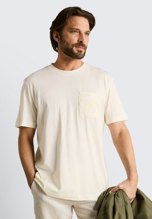 Mężczyzna z brodą, ubrany w kremową koszulę z krótkim rękawem i kieszenią na piersi, trzymający oliwkowozieloną kurtkę w prawej ręce na jednolitym tle.