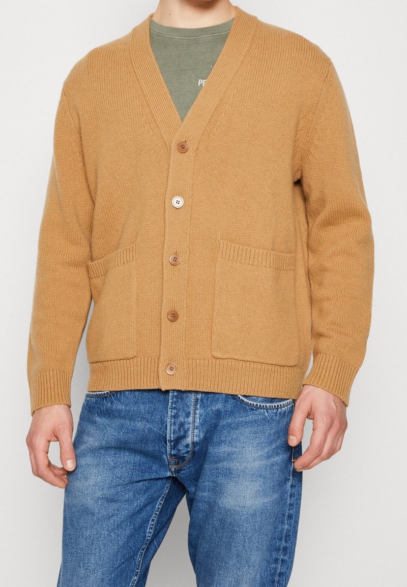 Cárdigan beige hecho de tejido de punto texturizado con escote en v, que presenta tres botones marrones, dos bolsillos delanteros y puños acanalados.