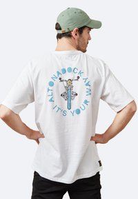 Camiseta blanca de algodón con un diseño gráfico de una bicicleta en el centro, rodeada de texto azul que dice "ALTONADOCK AVM IT'S YOUR."