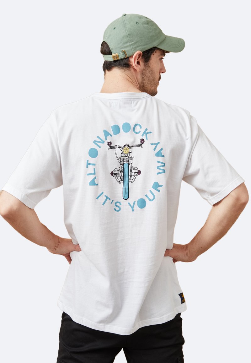 Camiseta blanca de algodón con un diseño gráfico de una bicicleta en el centro, rodeada de texto azul que dice "ALTONADOCK AVM IT'S YOUR."