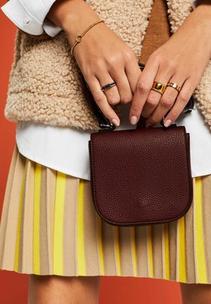 Handen met gouden en marineblauwe ringen houden een kleine, getextureerde donkerbruine leren handtas vast boven een gele geplooide rok met strepen.