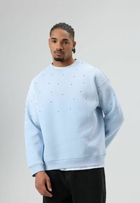Sweat-shirt bleu clair à coupe décontractée, orné de petits pois noirs, avec des poignets côtelés et un col rond. Porté avec un pantalon foncé.