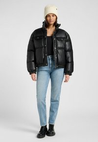 Chaqueta de plumas negra con botones a presión, puños elásticos y dos bolsillos delanteros. Llevada sobre una camiseta negra, combinada con jeans de color azul claro y botas negras.