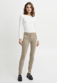 Pantalon beige slim avec des détails de boutonnage à l'avant et des poches latérales, associé à un haut blanc à manches longues. Des mocassins noirs complètent la tenue.