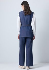 Gilet en denim avec taille ceinturée, porté par-dessus une blouse blanche à manches longues. Assorti à un pantalon en denim ample et des baskets blanches.