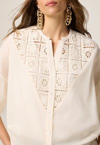 Blusa crema con inserto in pizzo a motivi geometrici, chiusura con bottoni discreta e vestibilità rilassata, che mette in evidenza texture e design intricati.