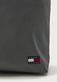 Γκρι υφασμάτινη τσάντα με υφή, διαθέτει ορθογώνια ετικέτα λογότυπου σε ναυτικό μπλε, λευκό και κόκκινο χρώμα που απεικονίζει την ένδειξη «TOMMY JEANS». 