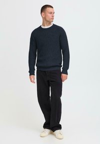 Man wearing dark blue knit sweater, black jeans, and white sneakers, standing and looking to his right against plain background.