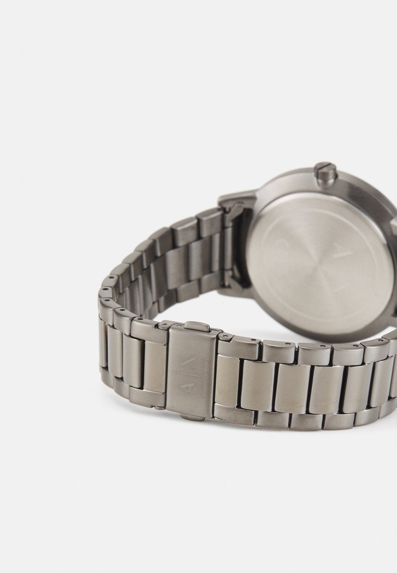 Reloj de acero inoxidable plateado con una caja redonda y suave, pulsera de eslabones cónicos y logo grabado en el cierre. Diseño minimalista, acabado mate.