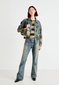 Denim jakna z cvetličnim vzorcem, prugasta pletena majica, svetlo modri široki kavbojke in črne čevlje s koničasto špicjo, ki drži majhno olivno zeleno torbico.