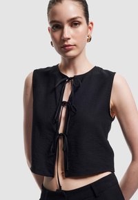 Zwarte cropped top met korte mouwen, textiel met een structuur, ronde halslijn en voorste knoopjes. Gecombineerd met zwarte hoge taillebroeken.