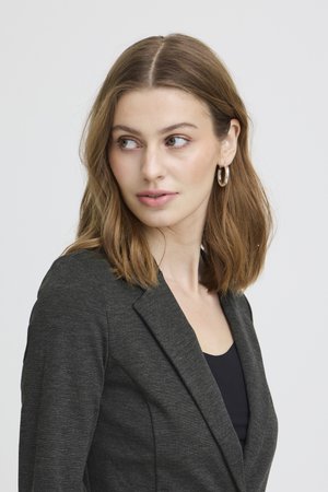 Mladá žena s vlasmi po plecia svetlo hnedej farby, ktorá má strieborné kruhové náušnice a tmavosivý blejzer cez čierne tričko, pozerá sa doľava.
