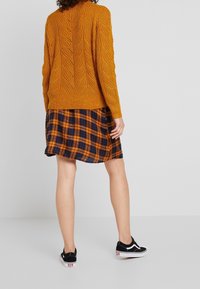 Pull orange tricoté avec motif en torsades, associé à une robe à carreaux bleu marine et orange. Des baskets noires basses complètent la tenue.