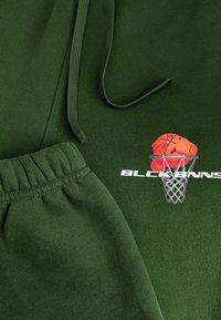 Groene sweatshirt en bijpassende joggingbroek, met een oranje basketbalafbeelding en de tekst "BLCK BNNS," met een gestructureerde afwerking en elastische tailleband.