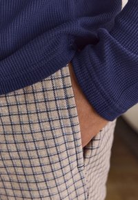 Navy ribbed sweater cuff alongside light beige pants featuring a blue check pattern, with a hand resting in the pants pocket.