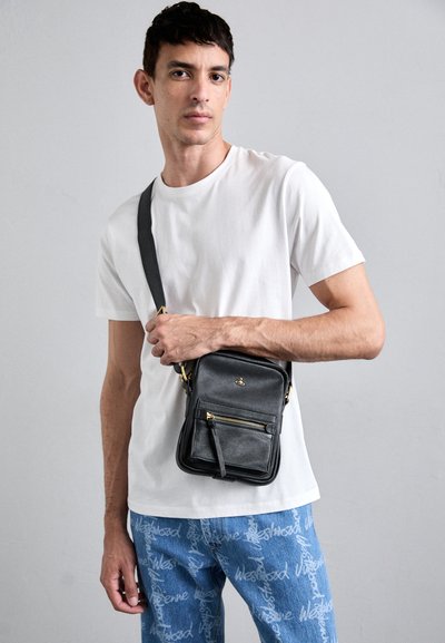 Černá kožená crossbody taška se zlatými akcenty, obdélníkový tvar a přední kapsa na zip. Kombinováno s bílým tričkem a vzorovanými džíny.