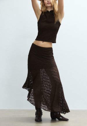 Nainen, joka pukeutunut hihattomaan mustaan pitsipaitaan ja vastaavaan pitkään pitsihameeseen, poseeraa nostetuin käsin, seisten vaalealla lattialla valkoista seinää vasten.