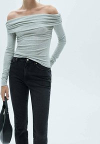 Blusa de manga larga gris claro, con un diseño sin hombros y detalles fruncidos, combinada con jeans ajustados negros. El conjunto se complementa con un bolso negro.