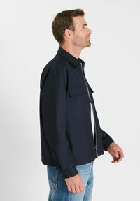 Marineblauw jack met rits en kraag, twee steekzakken aan de voorkant en lange mouwen. Gecombineerd met lichtblauwe spijkerbroek en wit T-shirt.