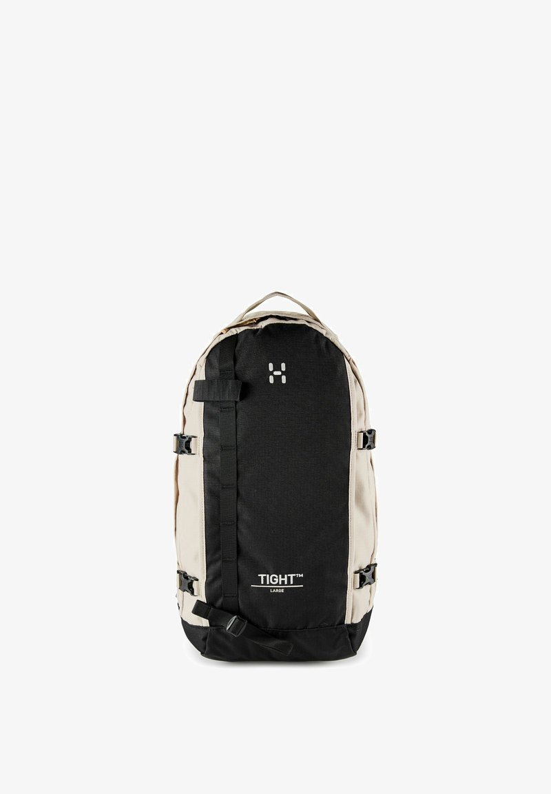 Grand sac à dos bicolore beige et noir avec poignée supérieure, sangles de compression latérales et logo « TIGHT » sur le panneau avant.