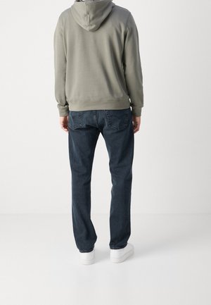 Homme portant un sweat à capuche gris, un jean bleu foncé et des baskets blanches, debout, dos à la caméra, sur un fond clair uni.