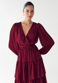 Bordowa sukienka kopertowa z długimi bufiastymi rękawami i warstwową spódnicą. Posiada dopasowaną talię z sznurkiem i miękką, teksturowaną tkaninę.