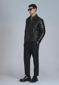 Schwarze gepolsterte Jacke mit glänzender Oberfläche, vier Fronttaschen und Knopfverschluss. Kombiniert mit schwarzen Hosen und Schnürschuhen.