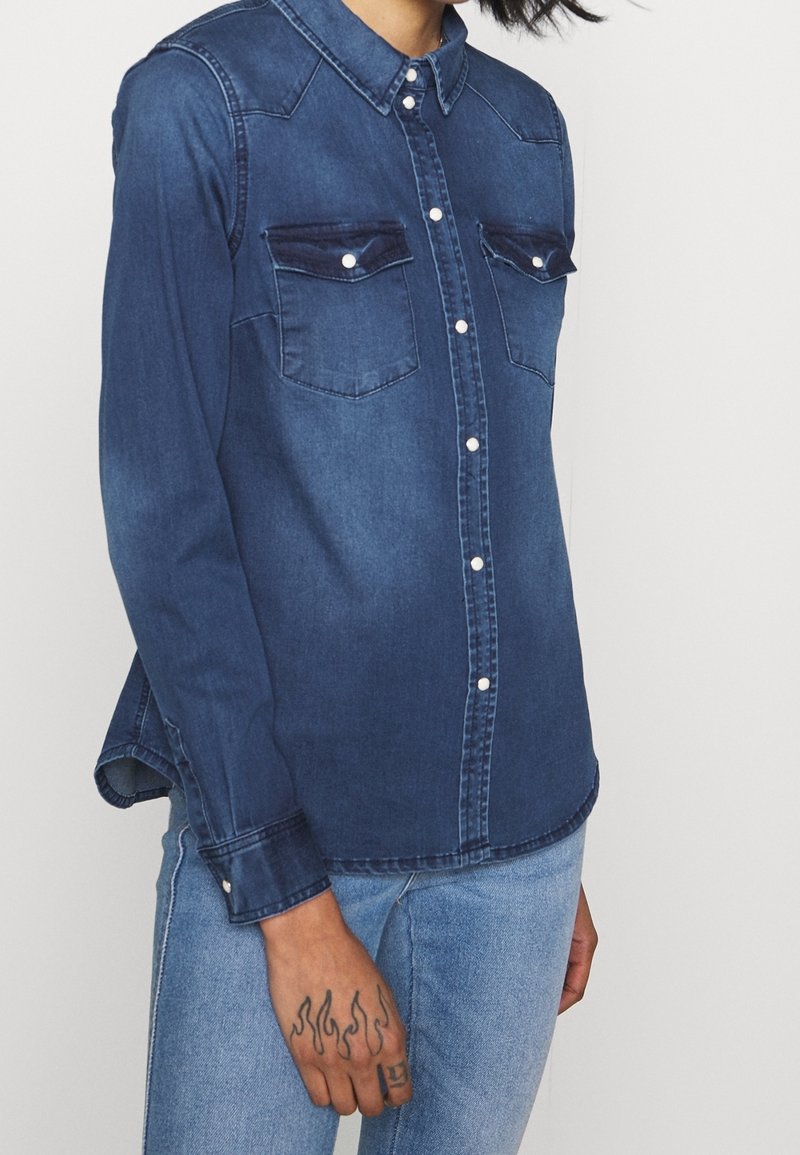 Personne portant une chemise en jean foncé à manches longues et un jean bleu clair, montrant un tatouage de flamme sur la main gauche.