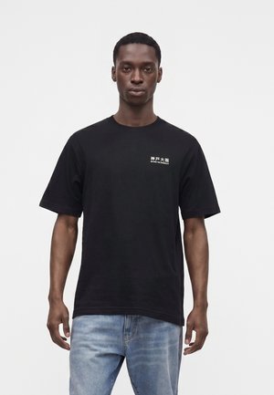 Homme portant un T-shirt noir avec des caractères asiatiques blancs et du texte, et un jean bleu clair, debout sur un fond blanc uni.