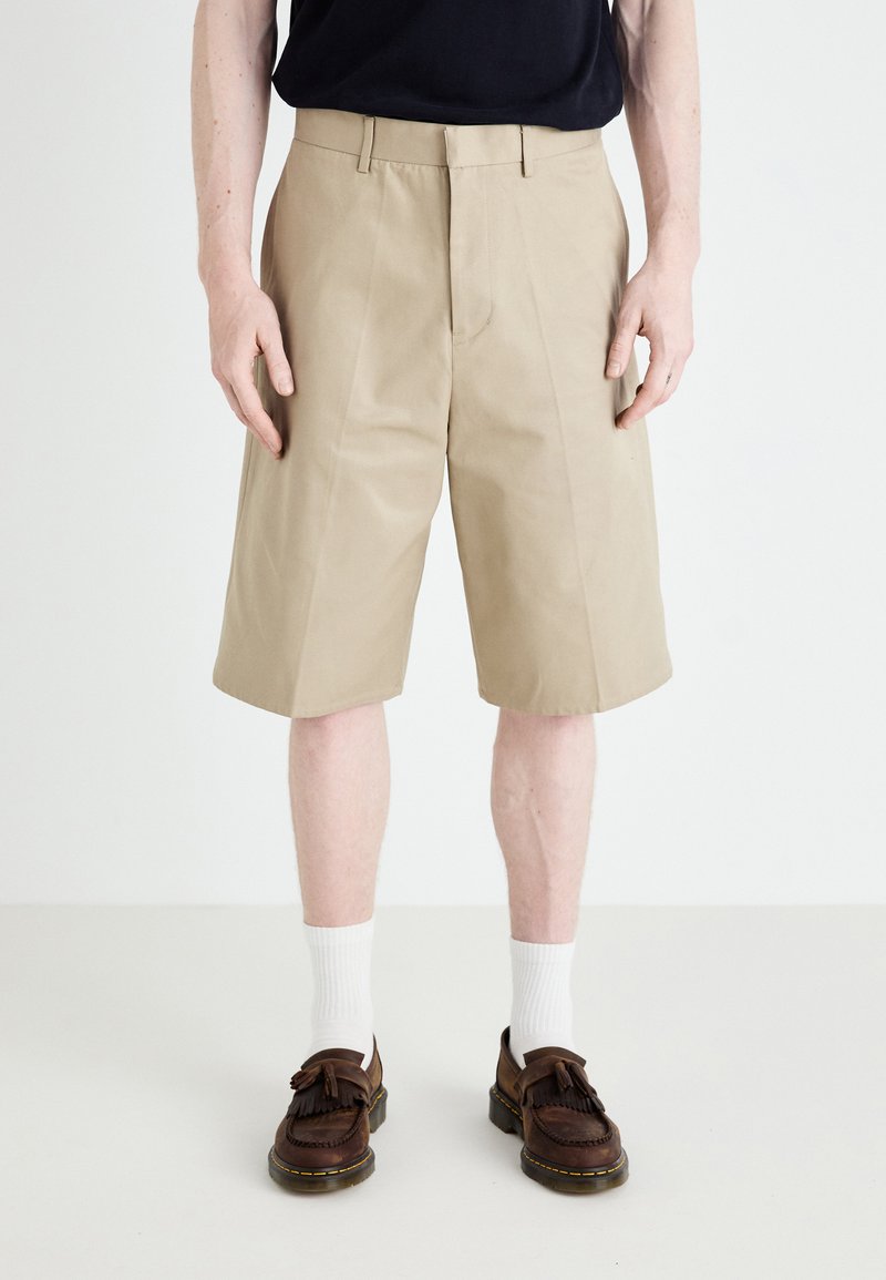 Pantalones cortos beige de mezcla de algodón, con corte recto, trabillas para cinturón y bolsillos laterales, combinados con mocasines marrones y calcetines blancos.