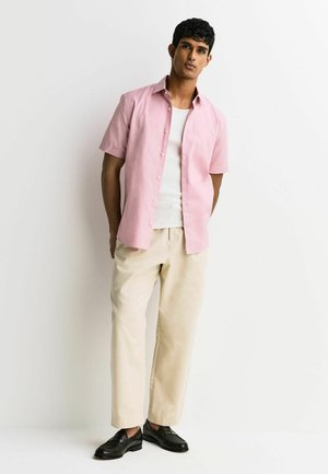 Mand, der står op ad en hvid væg iført lyserød kortærmet skjorte med knapper, hvid tanktop, beige bukser og sorte loafers.