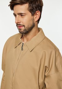 Beige Zip-Jacke mit umklappbarem Kragen aus strukturiertem Material. Sie verfügt über einen Reißverschluss und lockere Ärmel.