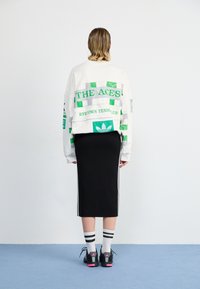 Biely oversized sweatshirt so zelenými a striebornými grafikami, skombinovaný s čiernou ceruzkovou sukňou, čiernymi teniskami a pruhovanými bielymi ponožkami.