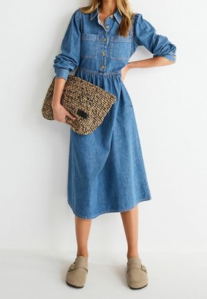 Frau mit einem langärmeligen Jeanskleid, die eine große geflochtene Clutch hält, kombiniert mit beigen Slippern.