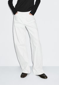 White wide-leg trousers with a smooth texture and flat front pockets, paired with a black long-sleeve top and dark shoes.