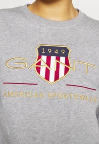 Sudadera gris con un escudo bordado con franjas rojas y blancas, texto dorado que dice "GANT" y "AMERICAN SPORTSWEAR", y detalle de 1949.