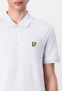Hellblaues Poloshirt aus strukturiertem Baumwollstoff, mit einer Drei-Knopf-Leiste und einem schwarz-gelben gestickten Vogel-Logo auf der Brust.