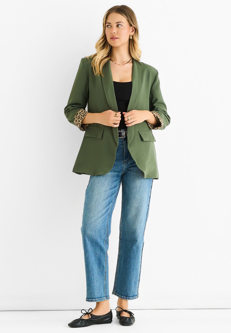 Blazer verde oliva con polsini a stampa leopardata, due tasche anteriori, abbinato a una maglia nera, jeans blu a gamba dritta e ballerine nere.