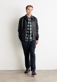Zwarte leren bomberjack over een geruit shirt, gecombineerd met donkere broeken en zwarte schoenen. Model staat tegen een neutrale achtergrond.